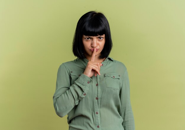 La giovane ragazza caucasica castana infastidita mette il dito sulla bocca che gesturing il segno silenzioso di silenzio isolato su fondo verde oliva con lo spazio della copia