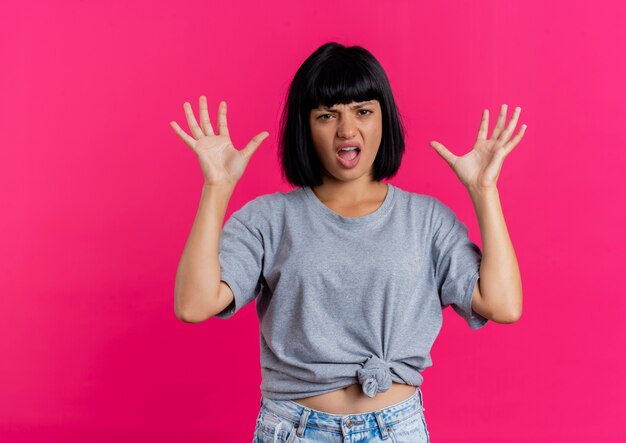 La giovane ragazza caucasica castana infastidita sta con le mani alzate che guarda l'obbiettivo isolato su fondo rosa con lo spazio della copia