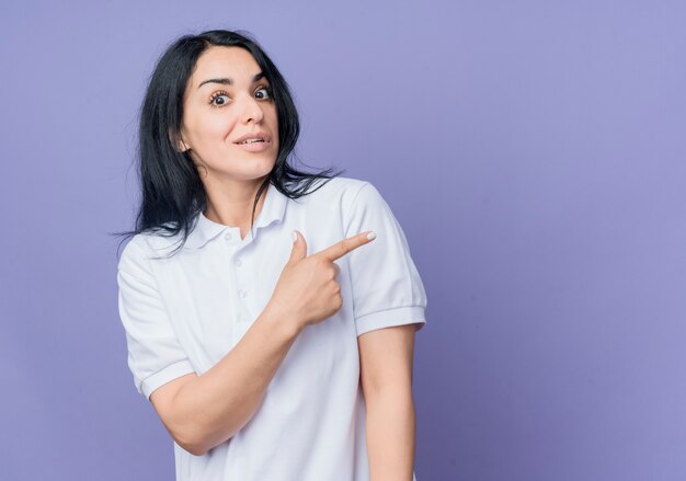 La giovane ragazza caucasica castana scioccata indica al lato che osserva isolato sulla parete viola