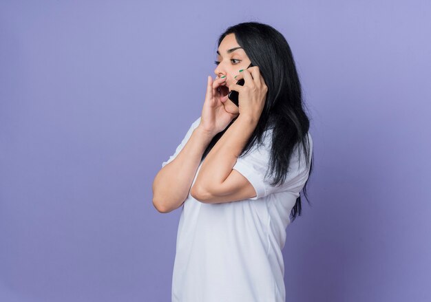 La giovane ragazza caucasica castana scioccata sta lateralmente parlando al telefono isolato sulla parete viola