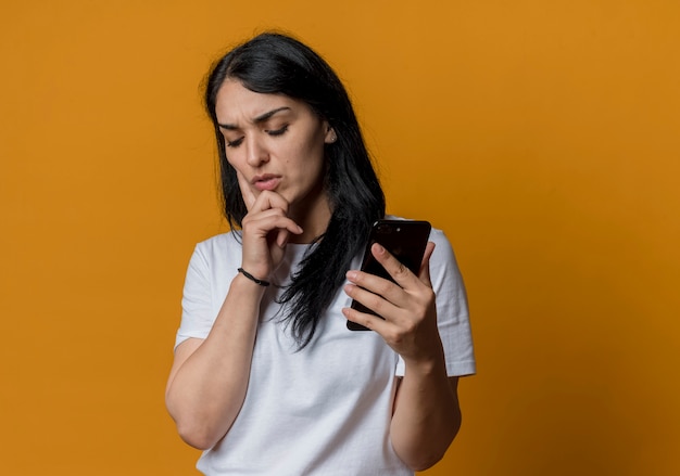 La giovane ragazza caucasica castana scontenta mette la mano sul telefono della tenuta del mento isolato sulla parete arancio