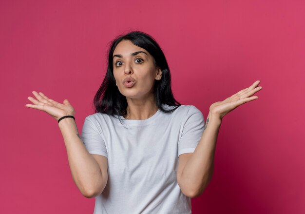 La giovane ragazza caucasica castana sorpresa sta con le mani aperte isolate sulla parete rosa