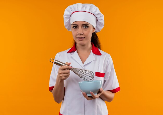 La giovane ragazza caucasica del cuoco in uniforme del cuoco unico tiene la ciotola e la frusta isolata su fondo arancio con lo spazio della copia