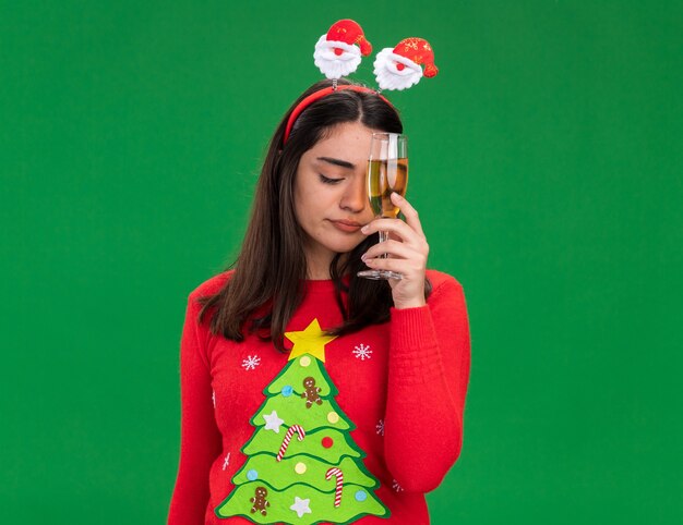 La giovane ragazza caucasica delusa con la fascia della Santa tiene il bicchiere di champagne isolato su priorità bassa verde con lo spazio della copia