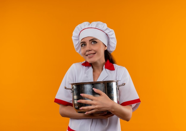 La giovane ragazza caucasica felice del cuoco in uniforme del cuoco unico abbraccia la pentola e osserva in su isolata sulla parete arancione con lo spazio della copia