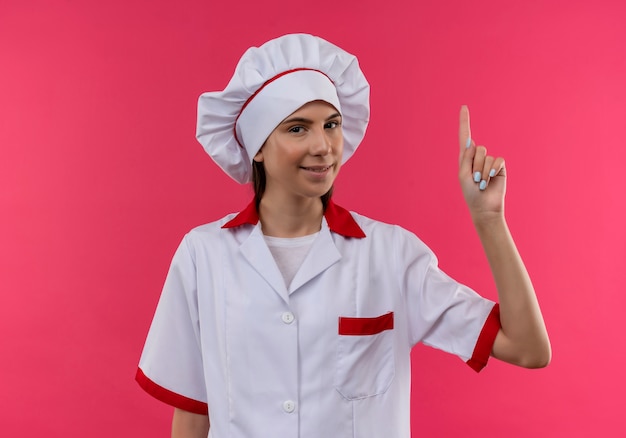 La giovane ragazza caucasica felice del cuoco in uniforme del cuoco unico indica in su isolato sullo spazio rosa con lo spazio della copia