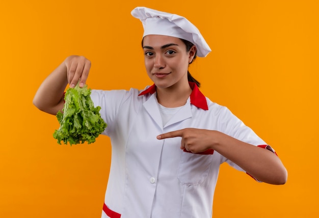 La giovane ragazza caucasica felice del cuoco in uniforme del cuoco unico tiene il dowm della parte superiore dell'insalata e punti con il dito isolato sulla parete arancione con lo spazio della copia