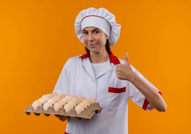 La giovane ragazza caucasica felice del cuoco in uniforme del cuoco unico tiene il lotto delle uova e dei pollici in su isolato sullo spazio arancione con lo spazio della copia