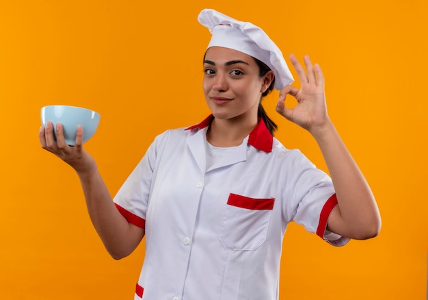 La giovane ragazza caucasica felice del cuoco in uniforme del cuoco unico tiene il segno giusto della mano di gesti e della ciotola isolato sulla parete arancio con lo spazio della copia