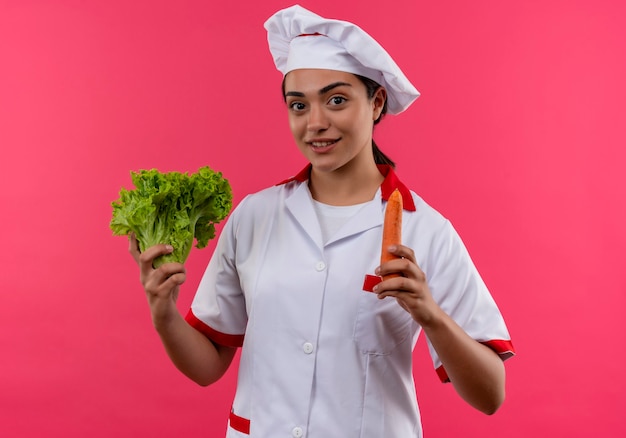 La giovane ragazza caucasica felice del cuoco in uniforme del cuoco unico tiene l'insalata e la carota isolate sulla parete rosa con lo spazio della copia