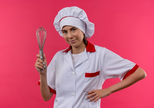 La giovane ragazza caucasica felice del cuoco in uniforme del cuoco unico tiene la frusta e mette la mano sulla vita sul colore rosa con lo spazio della copia