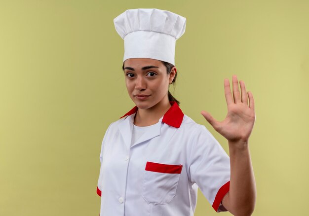 La giovane ragazza caucasica fiduciosa del cuoco in gesti uniformi del cuoco unico ferma il segno della mano isolato sulla parete verde con lo spazio della copia