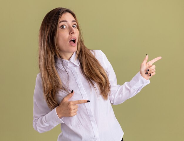 La giovane ragazza caucasica graziosa sorpresa indica a lato con due mani isolate sulla parete verde oliva con lo spazio della copia