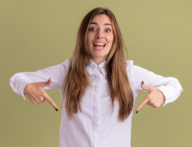 La giovane ragazza caucasica graziosa sorpresa indica giù con due mani su verde oliva