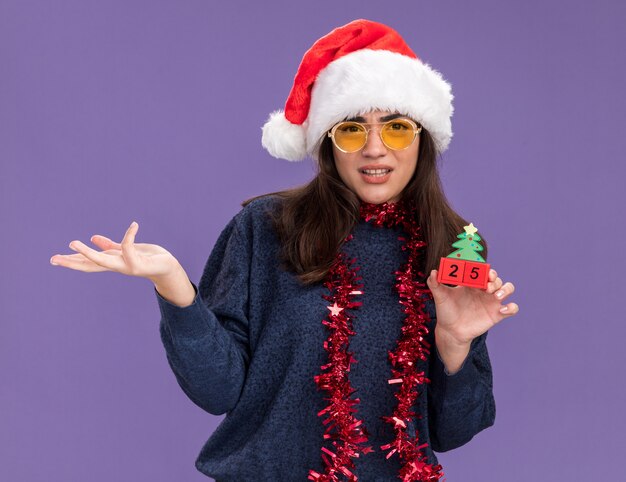 La giovane ragazza caucasica incosciente in occhiali da sole con cappello da Babbo Natale e ghirlanda intorno al collo tiene l'ornamento dell'albero di Natale isolato sulla parete viola con spazio di copia