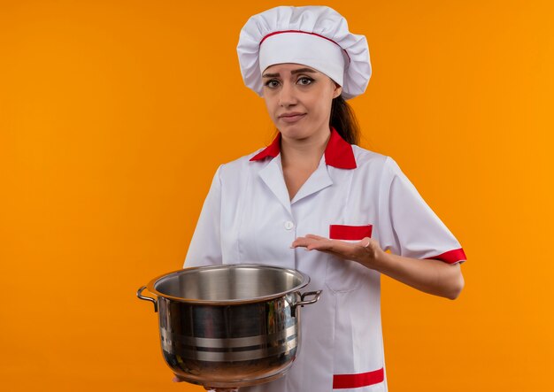 La giovane ragazza caucasica infastidita del cuoco in uniforme del cuoco unico tiene e punti alla pentola isolata su fondo arancio con lo spazio della copia