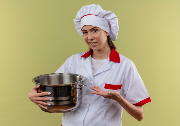 La giovane ragazza caucasica infastidita del cuoco in uniforme del cuoco unico tiene e punti alla pentola isolata su fondo verde con lo spazio della copia