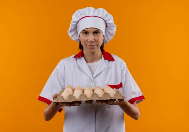 La giovane ragazza caucasica infastidita del cuoco in uniforme del cuoco unico tiene il lotto delle uova e guarda la macchina fotografica isolata sullo spazio arancione con lo spazio della copia