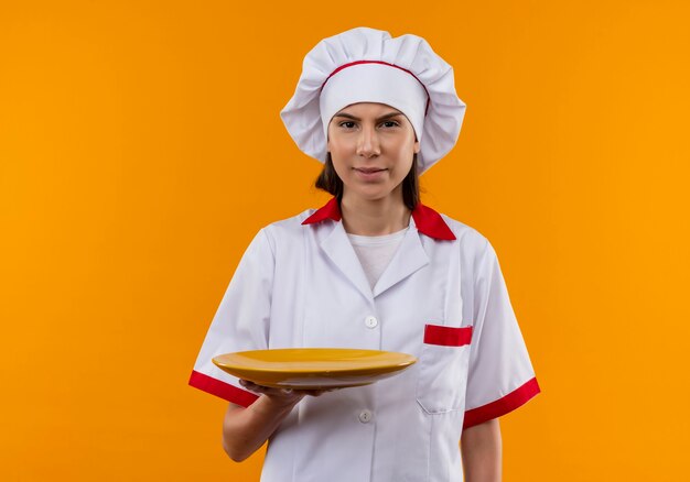 La giovane ragazza caucasica infastidita del cuoco in uniforme del cuoco unico tiene la zolla ed esamina la macchina fotografica isolata su fondo arancio con lo spazio della copia