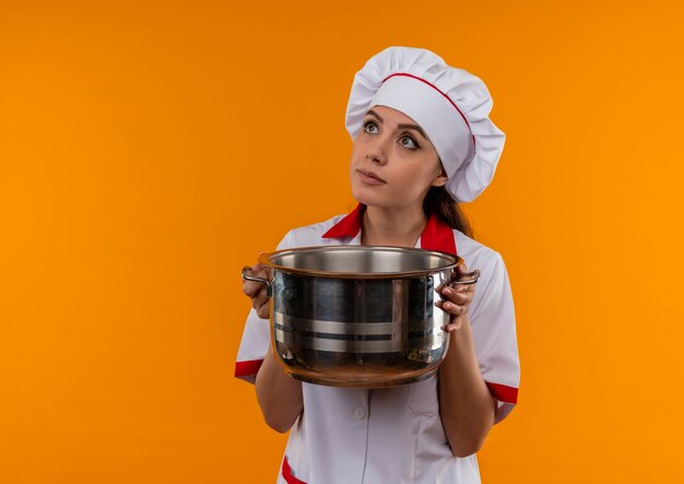 La giovane ragazza caucasica premurosa del cuoco in uniforme del cuoco unico tiene la pentola isolata sulla parete arancione con lo spazio della copia