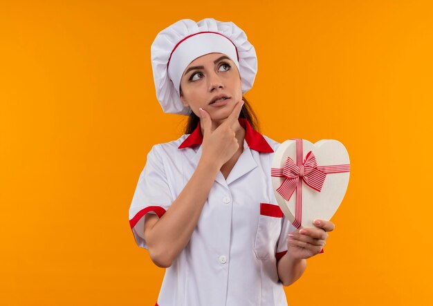 La giovane ragazza caucasica premurosa del cuoco in uniforme del cuoco unico tiene la scatola di forma del cuore e mette la mano sul mento isolata sulla parete arancione con lo spazio della copia