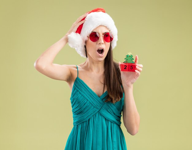 La giovane ragazza caucasica scioccata in occhiali da sole con il cappello della santa mette la mano sulla testa e tiene l'ornamento dell'albero di Natale che esamina il lato