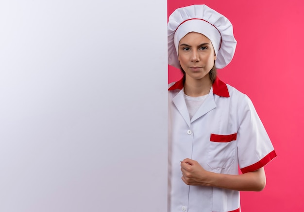 La giovane ragazza caucasica seria del cuoco in uniforme del cuoco unico sta dietro la parete bianca sul colore rosa con lo spazio della copia