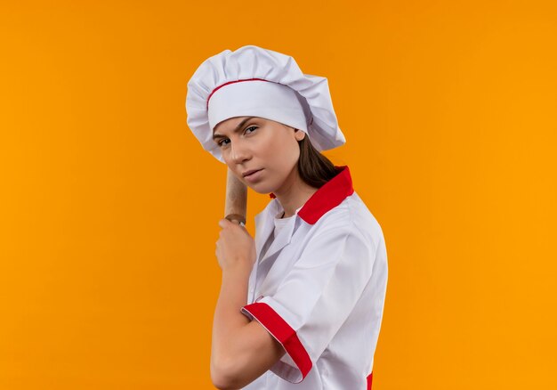 La giovane ragazza caucasica seria del cuoco in uniforme del cuoco unico sta lateralmente e tiene il mattarello dietro sull'arancia con lo spazio della copia