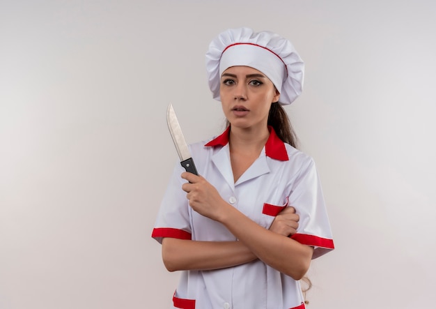 La giovane ragazza caucasica sicura del cuoco in uniforme del cuoco unico attraversa le braccia e tiene il coltello isolato su fondo bianco con lo spazio della copia
