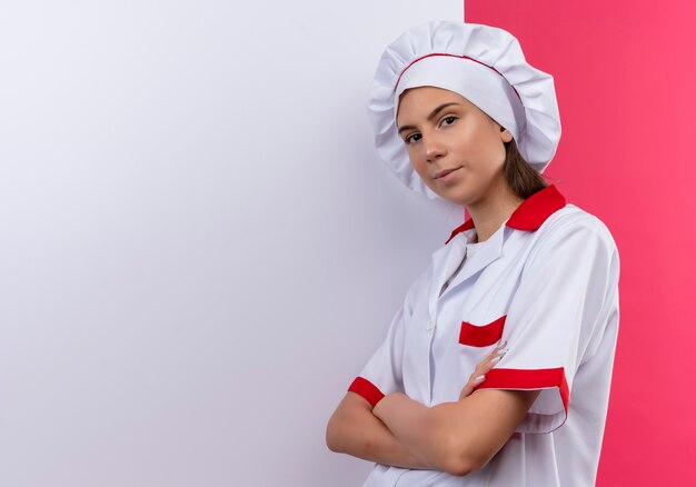 La giovane ragazza caucasica sicura del cuoco in uniforme del cuoco unico sta davanti alla parete bianca sul colore rosa con lo spazio della copia