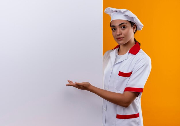 La giovane ragazza caucasica sicura del cuoco in uniforme del cuoco unico sta dietro la parete bianca e indica alla parete con la mano isolata sulla parete arancione con lo spazio della copia