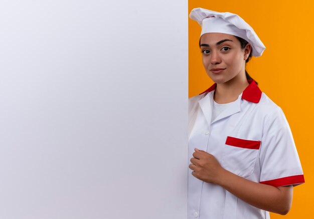 La giovane ragazza caucasica sicura del cuoco in uniforme del cuoco unico sta dietro la parete bianca e tiene il pugno isolato sulla parete arancione con lo spazio della copia