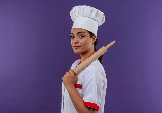 La giovane ragazza caucasica sicura del cuoco in uniforme del cuoco unico sta lateralmente e tiene il mattarello isolato sulla parete viola con lo spazio della copia