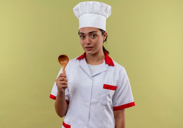 La giovane ragazza caucasica sicura del cuoco in uniforme del cuoco unico tiene il cucchiaio di legno isolato sulla parete verde con lo spazio della copia