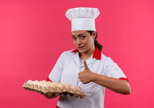 La giovane ragazza caucasica sicura del cuoco in uniforme del cuoco unico tiene il lotto delle uova e dei pollici in su isolati sulla parete rosa con lo spazio della copia