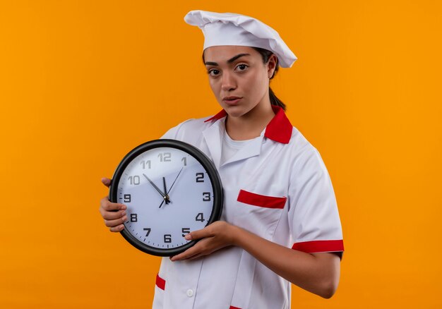 La giovane ragazza caucasica sicura del cuoco in uniforme del cuoco unico tiene l'orologio con entrambe le mani isolate sulla parete arancione