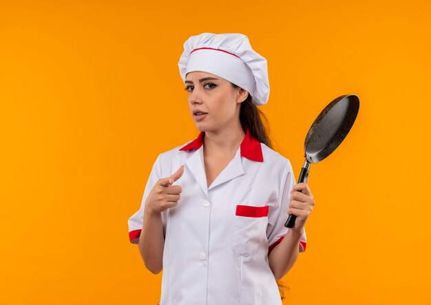 La giovane ragazza caucasica sicura del cuoco in uniforme del cuoco unico tiene la padella isolata sulla parete arancione con lo spazio della copia