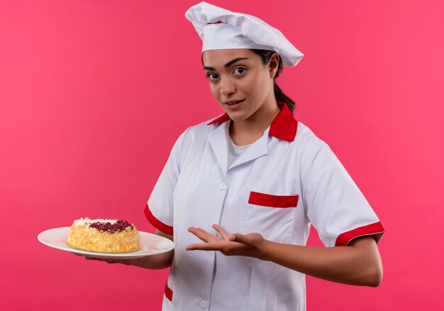 La giovane ragazza caucasica sicura del cuoco in uniforme del cuoco unico tiene la torta sul piatto e sui punti con la mano isolata sulla parete rosa con lo spazio della copia