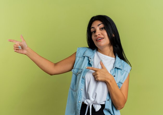 La giovane ragazza caucasica sicura indica a lato con due mani isolate su fondo verde oliva con lo spazio della copia