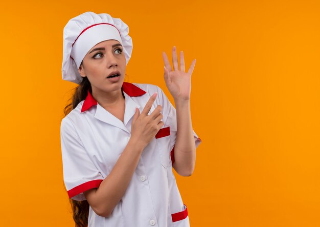 La giovane ragazza caucasica sorpresa del cuoco in uniforme del cuoco unico osserva e indica il lato isolato sulla parete arancione con lo spazio della copia