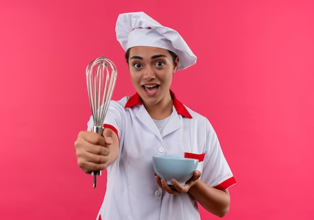 La giovane ragazza caucasica sorpresa del cuoco in uniforme del cuoco unico tiene la ciotola e tiene la frusta fuori isolata sulla parete rosa con lo spazio della copia