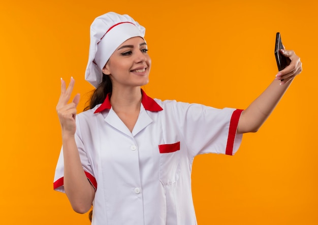La giovane ragazza caucasica sorridente del cuoco in uniforme del cuoco unico esamina il segno della mano di vittoria di gesti e del telefono isolato sulla parete arancio con lo spazio della copia
