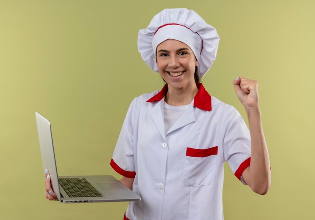 La giovane ragazza caucasica sorridente del cuoco in uniforme del cuoco unico tiene il computer portatile e solleva il pugno isolato su fondo verde con lo spazio della copia