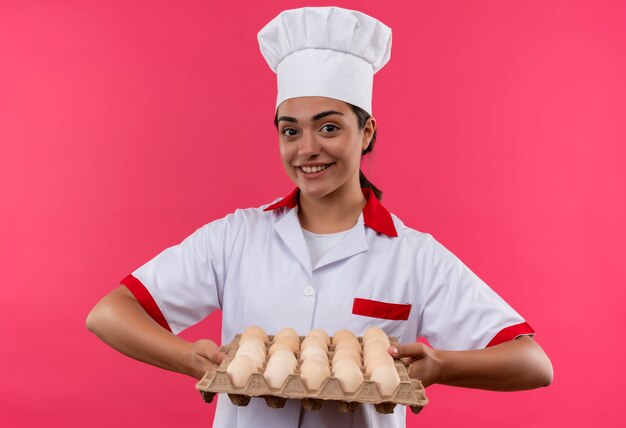 La giovane ragazza caucasica sorridente del cuoco in uniforme del cuoco unico tiene il lotto delle uova isolate sulla parete rosa con lo spazio della copia