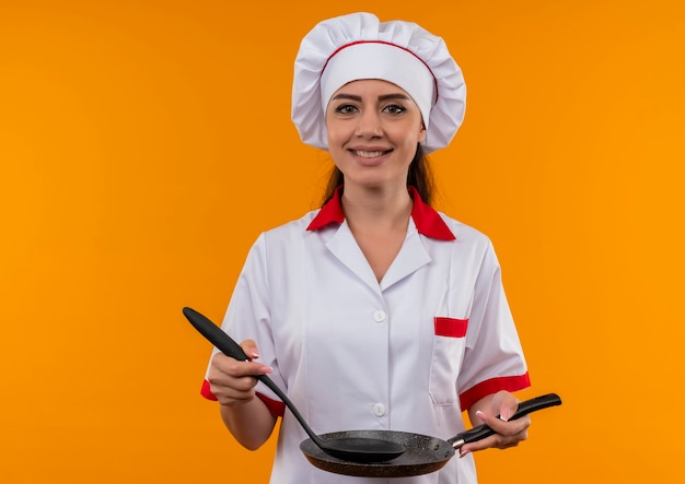 La giovane ragazza caucasica sorridente del cuoco in uniforme del cuoco unico tiene la padella e la spatola isolate sulla parete arancione con lo spazio della copia