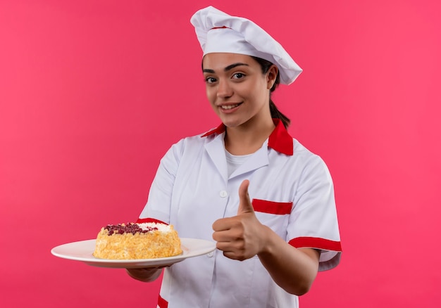 La giovane ragazza caucasica sorridente del cuoco in uniforme del cuoco unico tiene la torta sul piatto e sui pollici in su isolato sulla parete rosa con lo spazio della copia