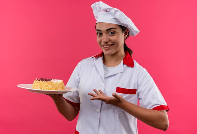 La giovane ragazza caucasica sorridente del cuoco in uniforme del cuoco unico tiene la torta sulla zolla e sui punti con la mano isolata sulla parete rosa con lo spazio della copia