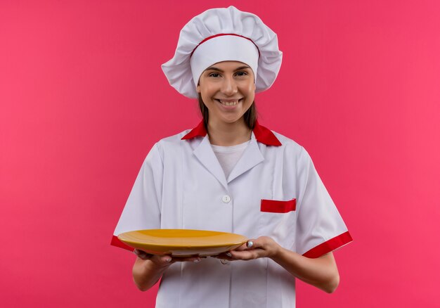 La giovane ragazza caucasica sorridente del cuoco in uniforme del cuoco unico tiene la zolla isolata sullo spazio rosa con lo spazio della copia