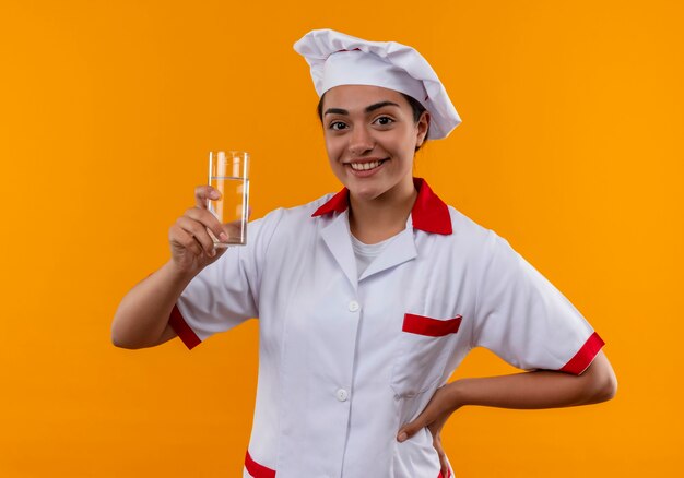 La giovane ragazza caucasica sorridente del cuoco in uniforme dello chef tiene il bicchiere d'acqua e mette la mano sulla vita isolata sulla parete arancione