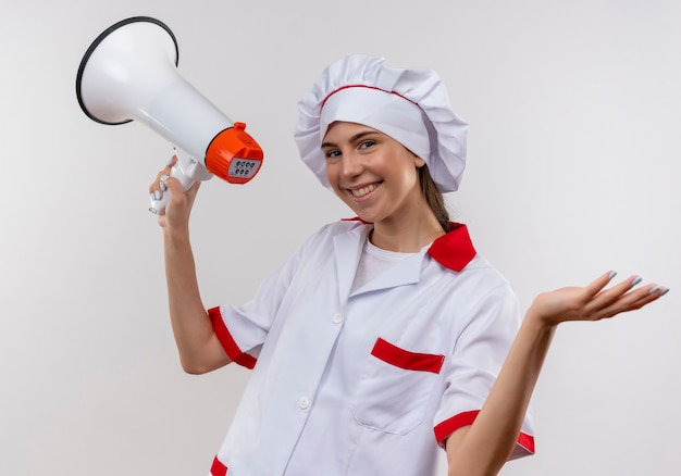 La giovane ragazza caucasica sorridente del cuoco in uniforme dello chef tiene l'altoparlante e tiene la mano aperta su bianco con lo spazio della copia